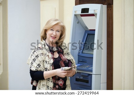 Mature blonde woman counting money near automated teller machine in shop