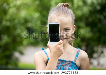 Happy little girl covers her face screen smartphone on a background of summer park