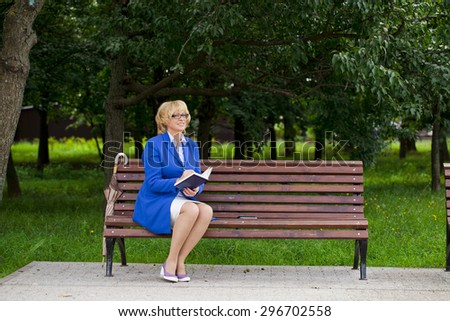 Mature business blonde woman in a jacket with diary sitting on a bench in a summer park