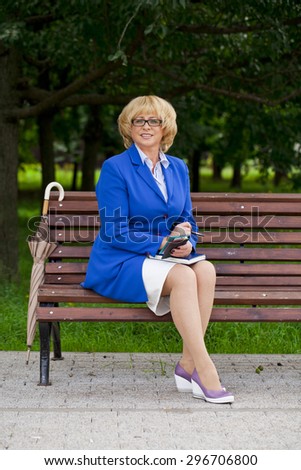 Elderly business woman in jacket sittin on bench with daily log, outdoor summer park