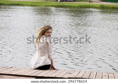 Young beautiful blonde woman in a white tunic sits on a wooden pier on the lake