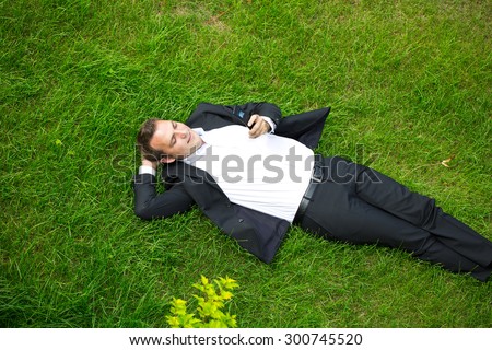 Cheerful young businessman calling by phone lying on green grass