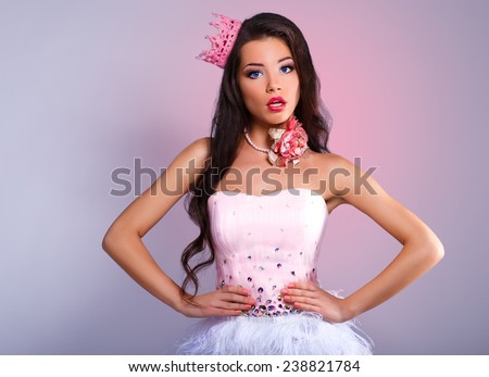 beautiful cheerful brunette girl in a pink dress and pink crown on his head. mini with sequins and feathers. long hair. gray background. surprise