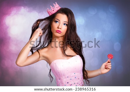 Beautiful cheerful brunette girl in a pink dress and pink crown on his head holding a lollipop. mini with sequins and feathers. long hair. gray background. surprise