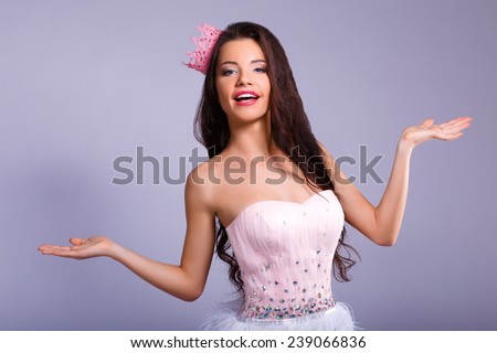 Beautiful cheerful brunette girl in a pink dress and pink crown on his head shows that a hands. mini with sequins and feathers. long hair.