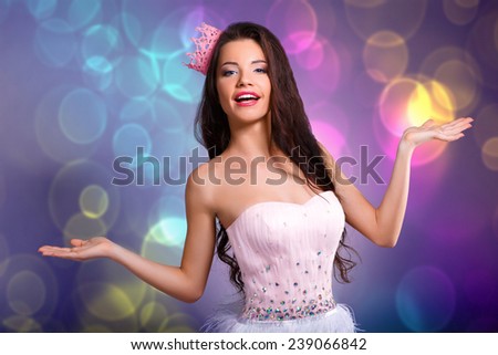 Beautiful cheerful brunette girl in a pink dress and pink crown on his head shows that a hands. mini with sequins and feathers. long hair.colorful background