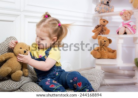 Little girl holding a teddy bear. Little girl in denim overalls sitting on cushions surrounded by toys