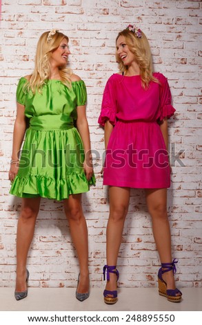 two fashion girls in bright dresses standing near the wall and look at each other.