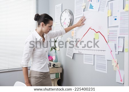 Female office worker bent over with head leaning to a wall and negative business chart.