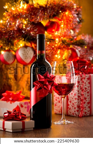 Wine bottle gift and wine glass filled with red wine, christmas gift boxes and tree on background.