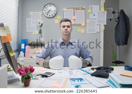Businessman at workplace wearing boxing gloves with fists on desktop and confident expression.
