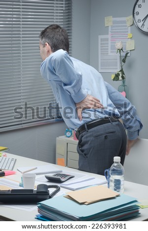 Tired businessman leaning to his desk and touching his painful lower back.