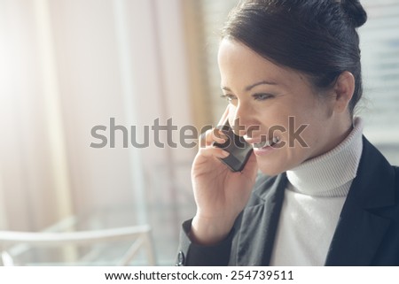 Attractive smiling woman having a phone call with her smart phone next to a window