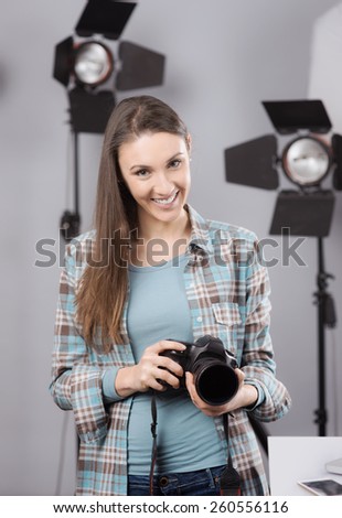 Young female photographer posing in her professional studio, holding a digital camera with lighting equipment on background