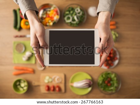 Hands holding a digital touch screen tablet with fresh vegetables and kitchen utensils on background, top view