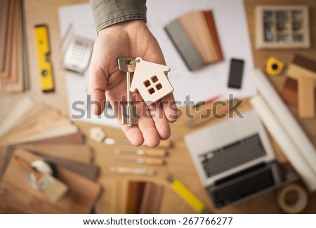 Real estate agent handing over a house key, desktop with tools, wood swatches and computer on background, top view