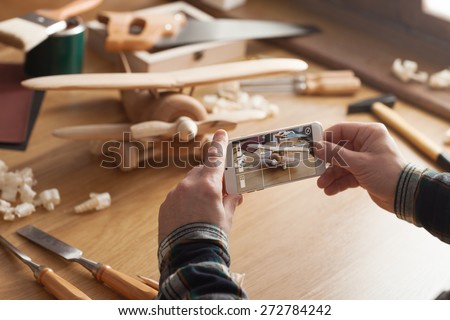Man photographing his handmade wooden toy airplane with a smart phone on a work table