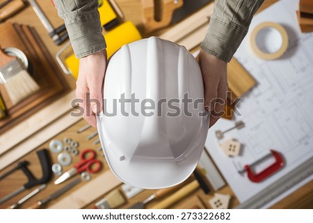 Hands holding a protective safety helmet against a work table with hardware and construction tools, top view