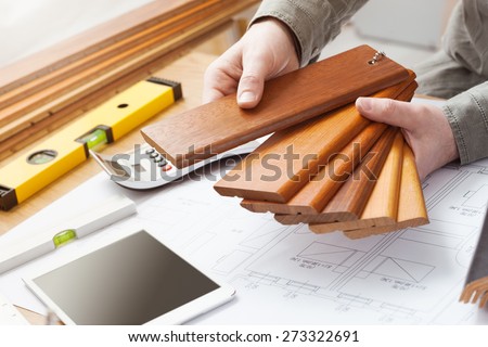 Professional interior designer holding wood swatches for baseboard and skirting, hands close up with desktop, house blueprint, tools and tablet
