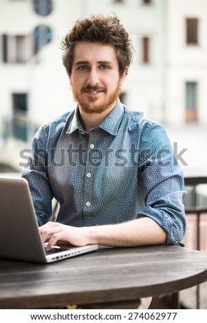 Smiling hipster young man working on laptop on a bar outdoor table
