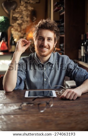 Cool hipster sitting at bar table and listening to music with his tablet using earphones