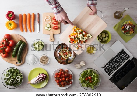 Man\'s hands cooking at home and chopping fresh vegetables on a cutting board, kitchen tools and food ingredients all around, top view