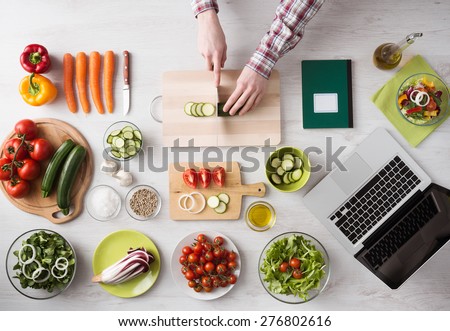 Man\'s hands cooking at home and chopping fresh vegetables on a cutting board, kitchen tools and food ingredients all around, top view