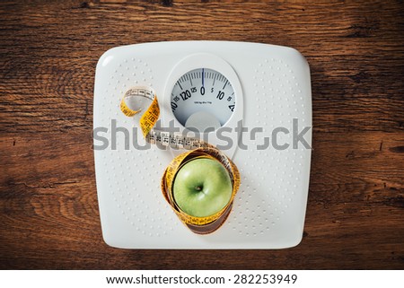 Green apple wrapped in a tape measure on a white scale, wooden surface on background, dieting and weight loss concept
