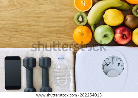 Fitness and weight loss concept, dumbbells, white scale, towel, fruit and mobile phone on a wooden table, top view