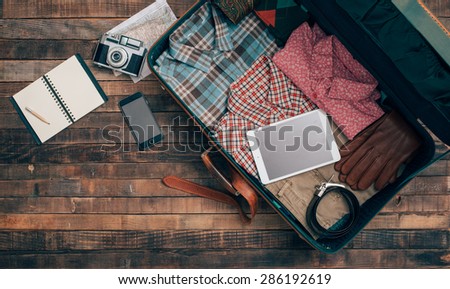 Vintage hipster traveler packing, open suitcase on a wooden table with clothing, camera and mobile phone, top view