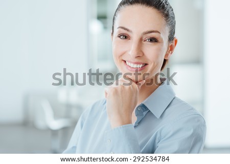 Pretty young woman posing with hand on chin and smiling at camera, room interior on background