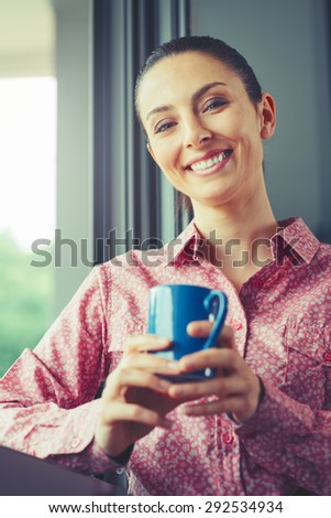 Woman having a relaxing coffee break at window, she is smiling and holding a mug