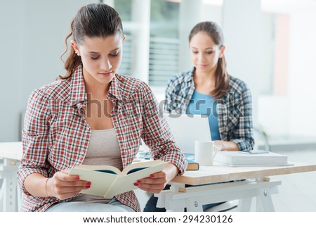 Cute girl at home reading a book and studying, her friend is sitting at desk using a laptop