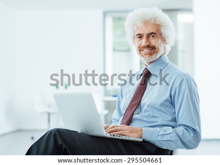 Smiling confident businessman looking at camera and typing on a laptop on his lap, he is sitting on an office chair, room interior on background