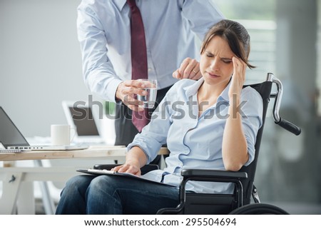 Business woman in wheelchair having an headache at office, her collegue is giving her a glass of water and helping her