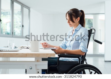 Confident disabled business woman in wheelchair working at office desk and checking paperwork