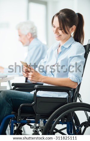Confident business woman in wheelchair working at office desk and texting with her mobile phone, disability and employment concept