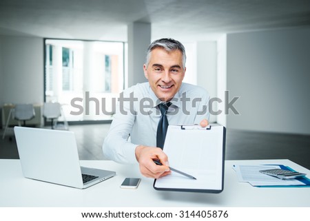 Confident businessman at desk handing a clipboard and a pen, contract signature and agreement concept