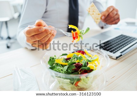 Businessman having a lunch break at desk, he is eating fresh salad and holding a cracker, unrecognizable person