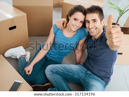Happy young couple thumbs up and smiling at camera, they are sitting on their new house floor surrounded by carton boxes