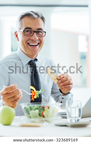 Smiling businessman sitting at office desk and having a lunch break, he is eating a salad bowl