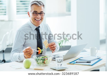 Smiling businessman sitting at office desk and having a lunch break, he is eating a salad bowl