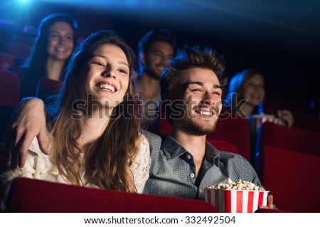 Young loving couple at the cinema watching a movie and smiling, people sitting on background