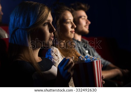 Teenager friends at the cinema watching a movie together and eating popcorn, beautiful girl on foreground, movies and entertainment concept