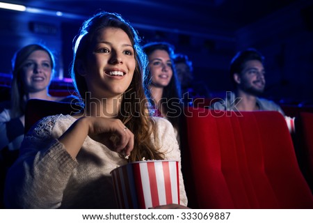 Young smiling woman watching a film in the movie theater and eating popcorn, entertainment and cinema concept