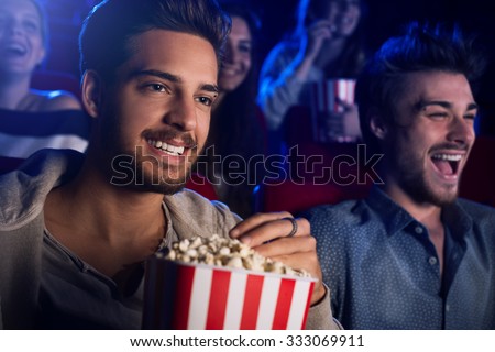 Young people sitting at the cinema, watching a movie and eating popcorn, two smiling men on foreground