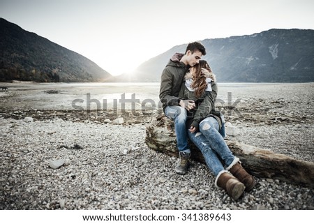 Young couple having a romantic date at the lake, they are hugging and sitting on a trunk on the beach, love and relationships concept