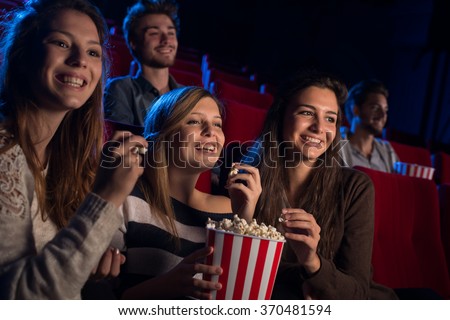 Smiling young girls sitting at the cinema, watching a film and eating popcorn, friendship and entertainment concept
