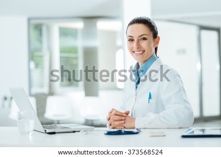Female doctor working at office desk and smiling at camera, office interior on background