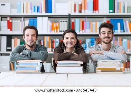 Young college students at the library studying together, they are smiling at camera and leaning on a pile of books